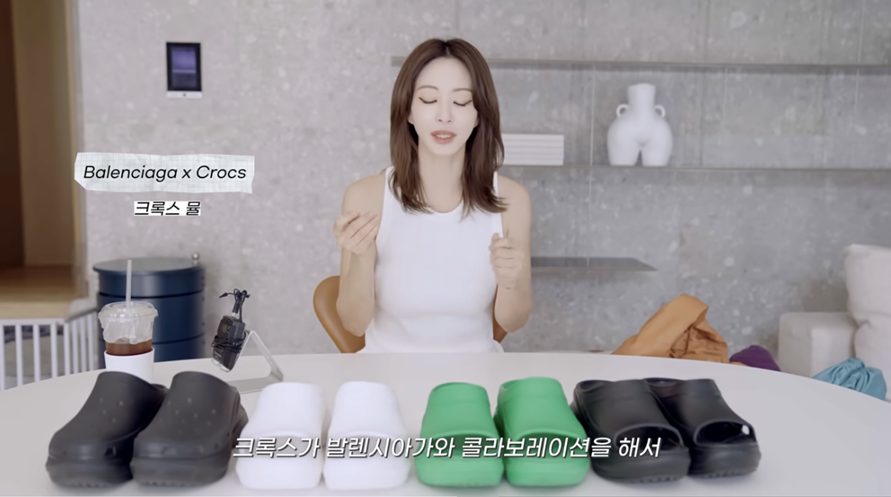 한예슬이 유튜브 영상에서 발렌시아가 크록스 뮬과 구찌 러버 슬라이드 샌들을 비교 소개하는 장면, 다양한 컬러의 샌들이 테이블 위에 진열된 모습