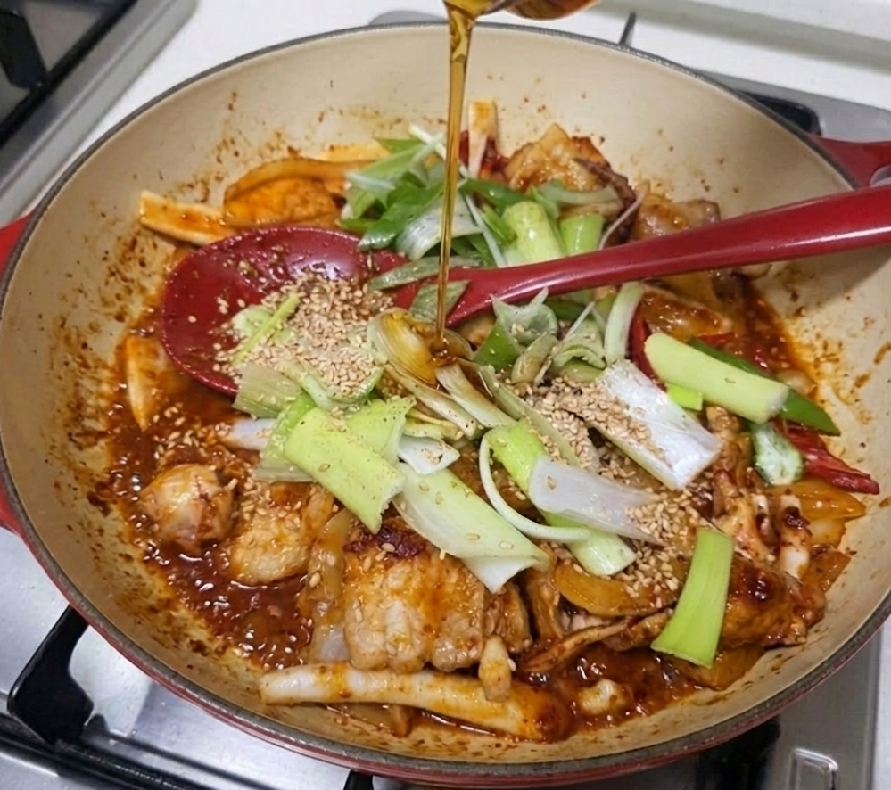 무쇠 팬에 담긴 오삼불고기 조리 과정. 빨간색 조리용 스푼이 놓여 있고, 위에서 참기름이 떨어지며 대파와 통깨가 뿌려진 먹음직스러운 모습.