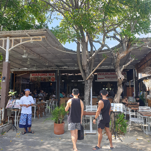 음식점 앞 큰 나무가 있다. 두 명의 고객이 들어가고 있고 입구에는 직원이 고객을 기다린다.