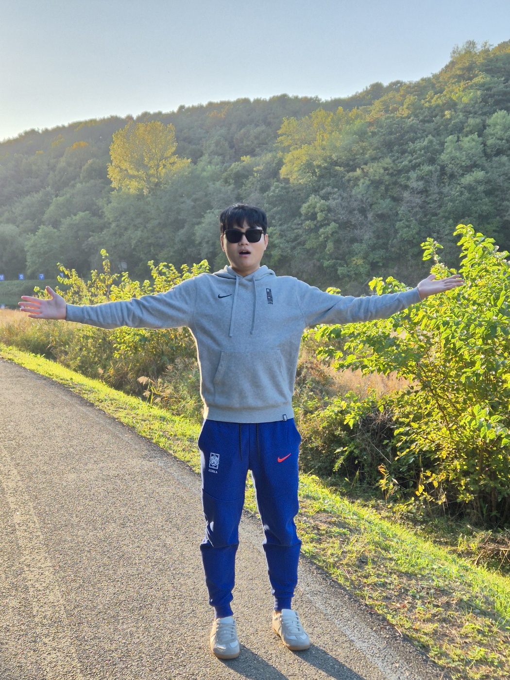 푸른 산을 배경으로 선글라스를 끼고 양팔을 벌려 자신감 있는 포즈를 취하고 있는 남성의 모습