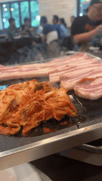 돌판 위에 삼겹살과 김치가 지글지글 익고 있는 장면. 뜨거운 연기 속 고소한 점심 한 끼의 시작