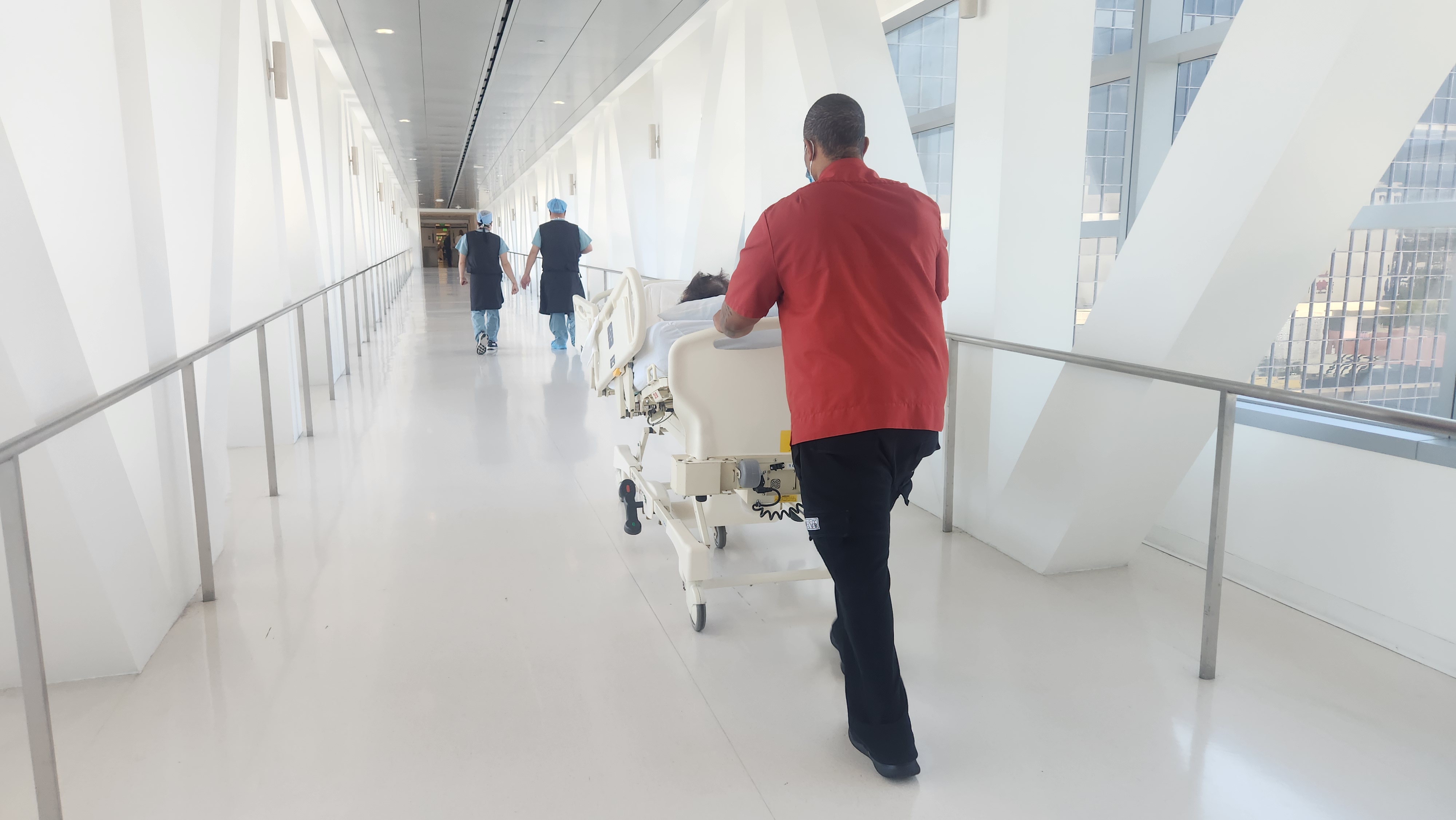 Cedars-Sinai 병원의 넓고 밝은 복도에서, 한 직원이 환자를 태운 침대를 밀고 수술실 방향으로 이동하는 모습 이미지