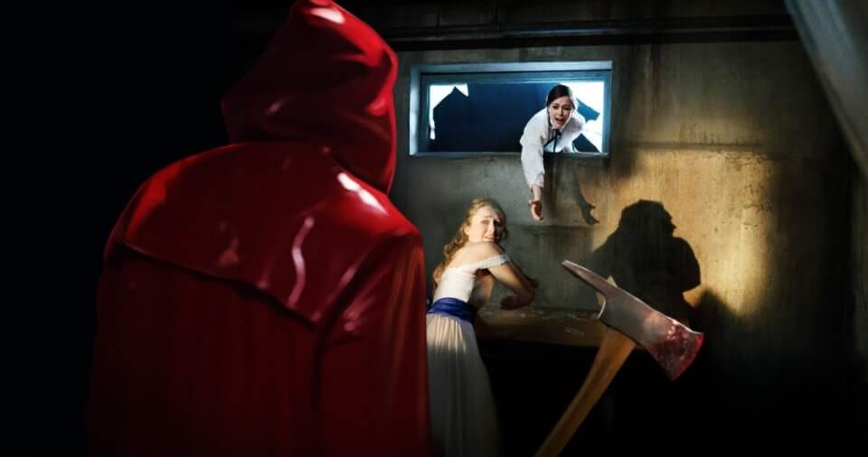 Fear Street: Prom Queen_붉은 망토를 입은 살인마가 피 묻은 도끼를 들고 있고, 프롬 드레스에 입은 젊은 여성이 겁에 질린 표정으로 도망치려 하고, 깨진 창문 너머에선 여성이 친구를 구하려고 손을 뻗고 있다.