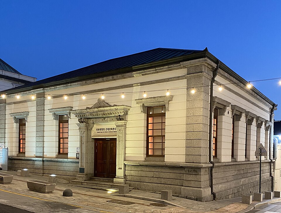 The former 18th Bank branch in Incheon, with a pale facade, a wooden doorway and tall windows beneath strings of warm lights and a blue evening sky.