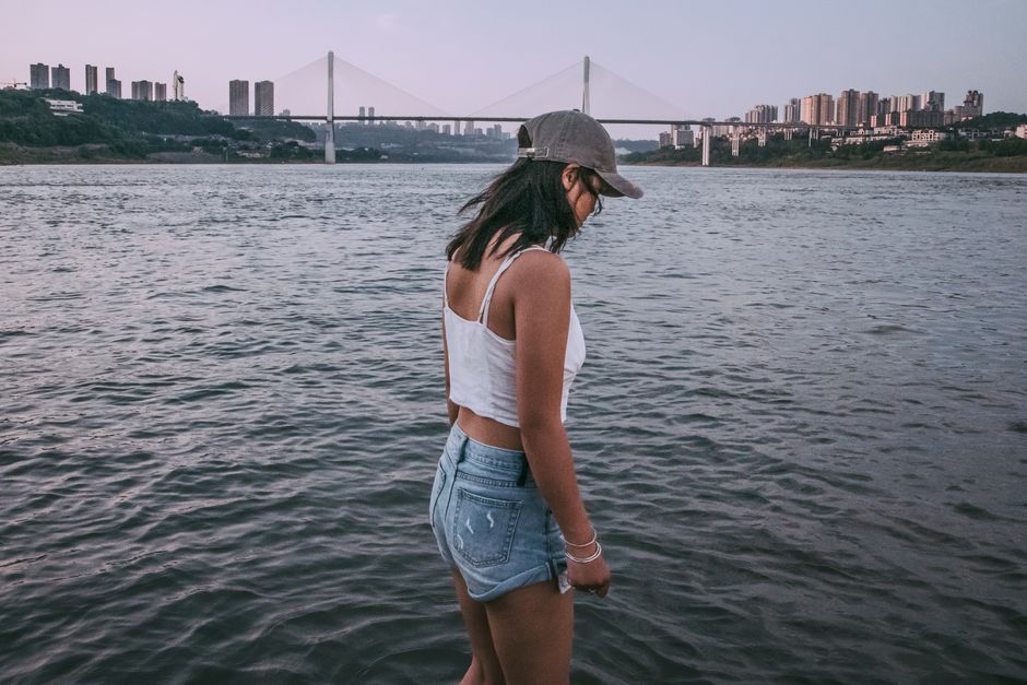 Young woman standing by a river with cityscape and bridge in background, summer vibe.