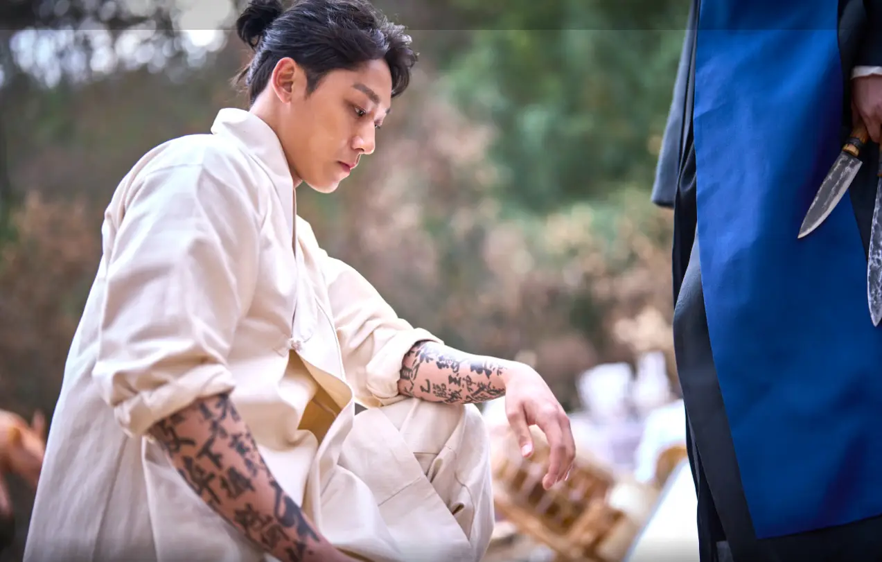 파묘- 흰 한복을 입고 팔에는 어지럽게 검은 한문글씨가 적힌채 쪼그리고 앉아 땅을 보고있는 이도현