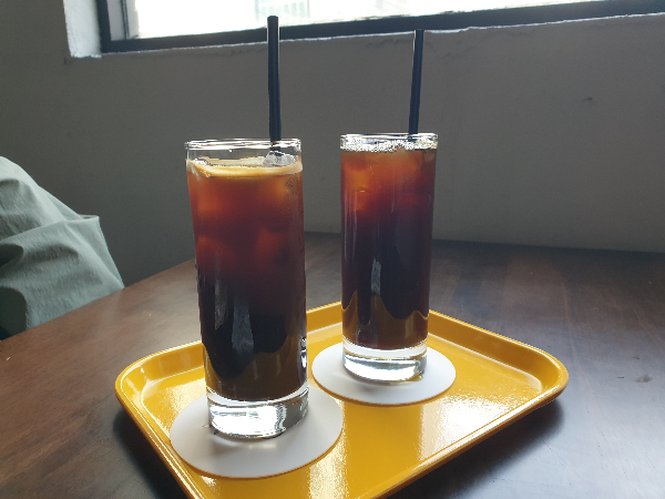 노란 쟁반 위에 아이스커피 2잔이 올려져 있다. 커피에는 검은색 빨대가 꽂혀 있다.