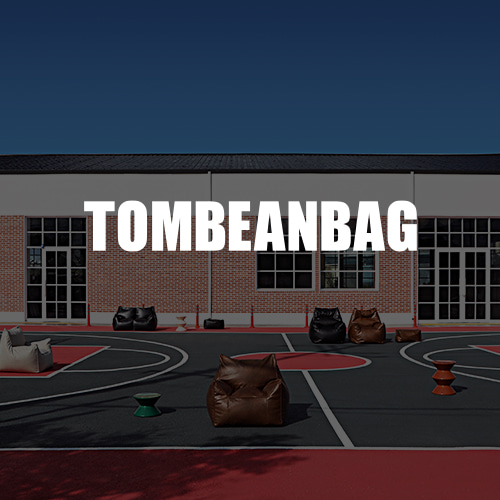 붉은 벽돌 건물 앞 농구 코트에 플래지어 톰빈백 빈백 소파 여러 점을 놓고 가운데에 TOMBEANBAG 글자를 얹은 이미지