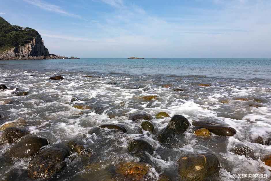 덕적도 능동자갈마당 해변의 몽돌과 바닷물. 크고 둥근 자갈들 위로 잔잔한 파도가 밀려들며 청아한 파도소리가 울리는 풍경. 뒤편으로 기암절벽과 푸른 바다가 조화를 이룬다