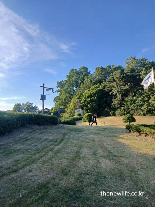 A man walks across the trimmed lawn of Children’s Grand Park, surrounded by dense trees and signage./ 울창한 나무와 표지판 사이를 걷는 서울 어린이대공원 잔디밭의 사람