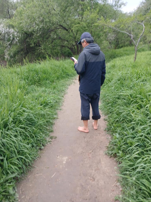 A man walking barefoot on a quiet dirt trail, looking at his phone-서울 여의도 샛강생태공원에서 흙길 위를 맨발로 걷으며 휴대폰을 보는 남성
