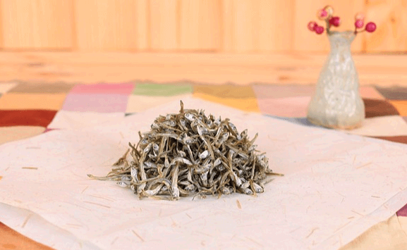 하얀 천 위에 회색의 마른 멸치들이 한가득 있다. 오른쪽엔 하얀 꽃병에 진분홍색 꽃이 꽂아있다.