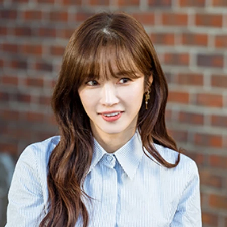 어느 벽돌 건물을 배경으로 하늘색 셔츠에 긴 갈색 웨이브 머리를 하고 있는 오늘도 사랑스럽개의 윤채아 캐릭터