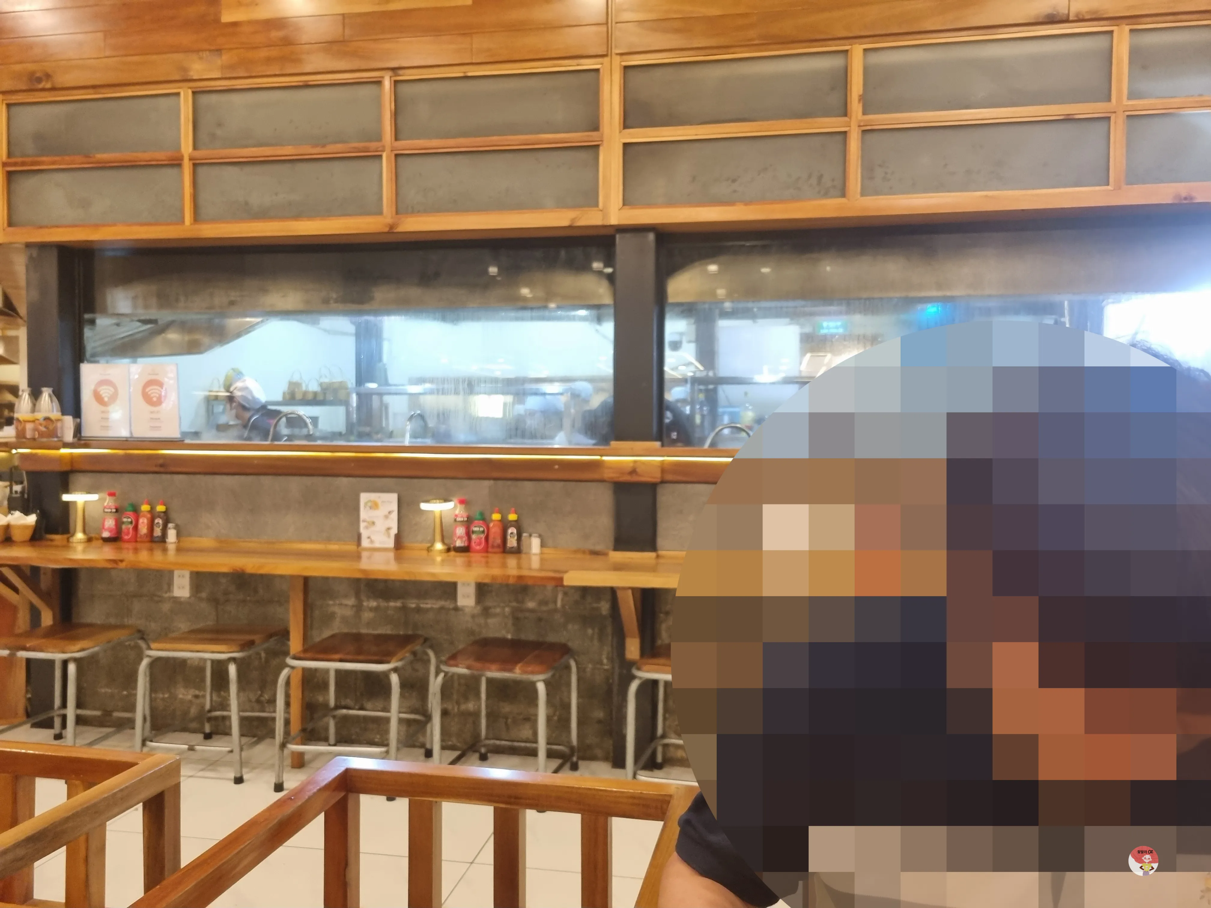 나무 인테리어의 식당 내부. 조리 공간이 유리창 너머로 보이고, 앞에는 좌석과 조명, 소스병이 있습니다.