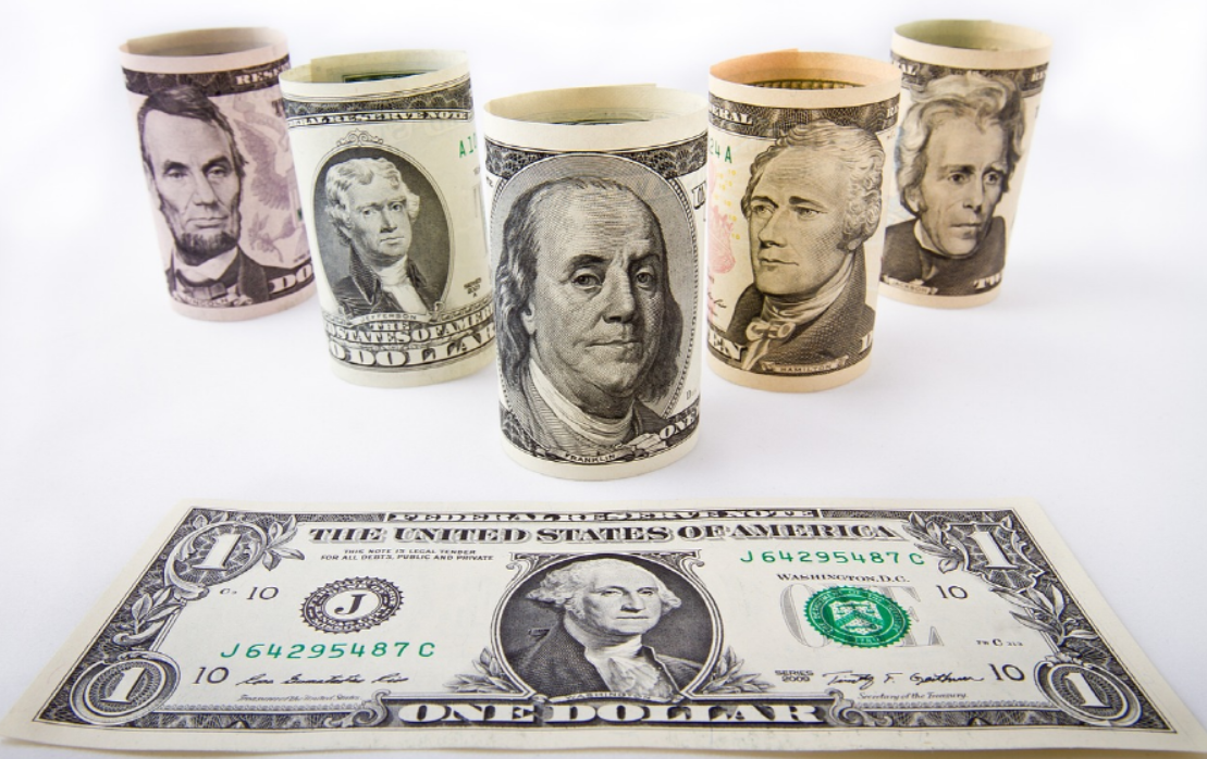 미국의 역대 대통령의 얼굴이 그려진 지폐 뭉치가 동그랗게 말려 있는데, 다섯 뭉치가 세워져 있다. 바닥에는 달러 지폐 한 장이 펼쳐져 있다.