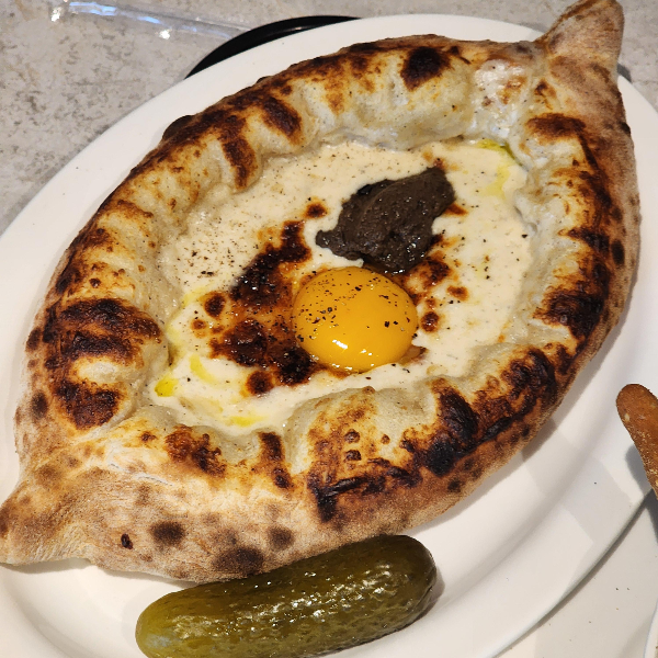 흰색 접시에 담긴 하차푸리(조지아 전통 빵). 바삭하게 구워진 빵 안에 치즈, 달걀 노른자, 그리고 트러플 페이스트가 올려져 있으며, 옆에는 피클이 함께 제공됨
