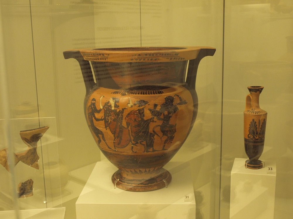 박물관 유리 진열장에 전시된 그리스 도기. 검은색으로 인물과 장식이 정교하게 그려진 대형 도자기가 중심에 놓여 있다.