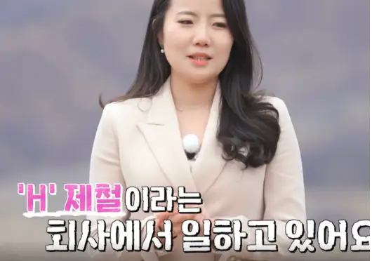 야산배경 흰정장입여자가 긴머리를 늘어뜨린채 말하는 모습 아래 분홍글싸'H'제철이라는 회사에서 일하고 있어요