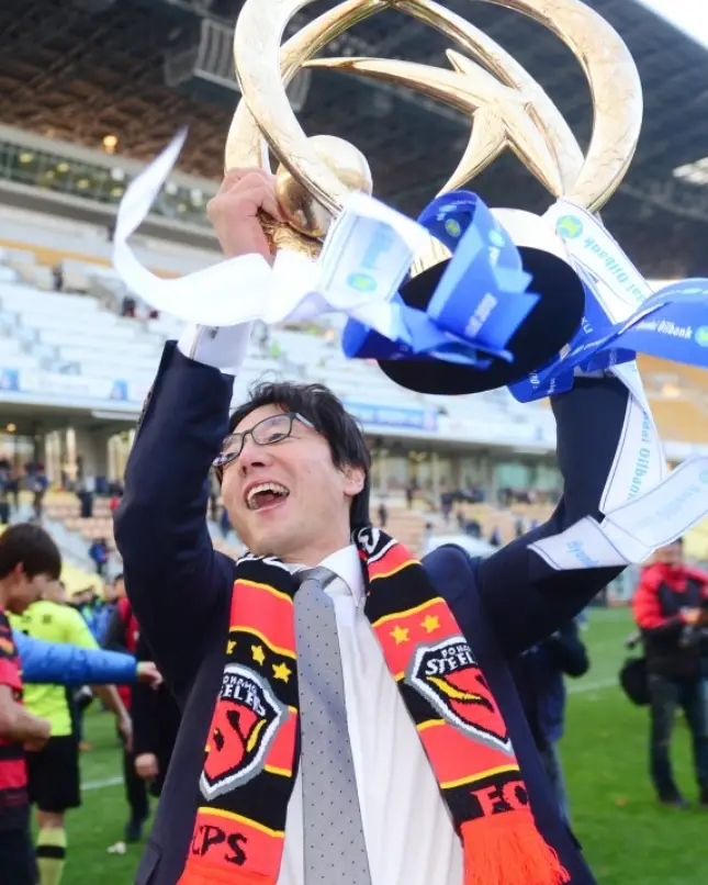 황선홍- 축구그라운드 뒤에는 취재진들이 빼곡히 서있고 파란 양복 흰와이셔츠 회색넥타이를 매고 목에는 축구구단의 목도리를 두른채 두손높이 우승트로피를 들고 기뻐하는 황선홍 감독