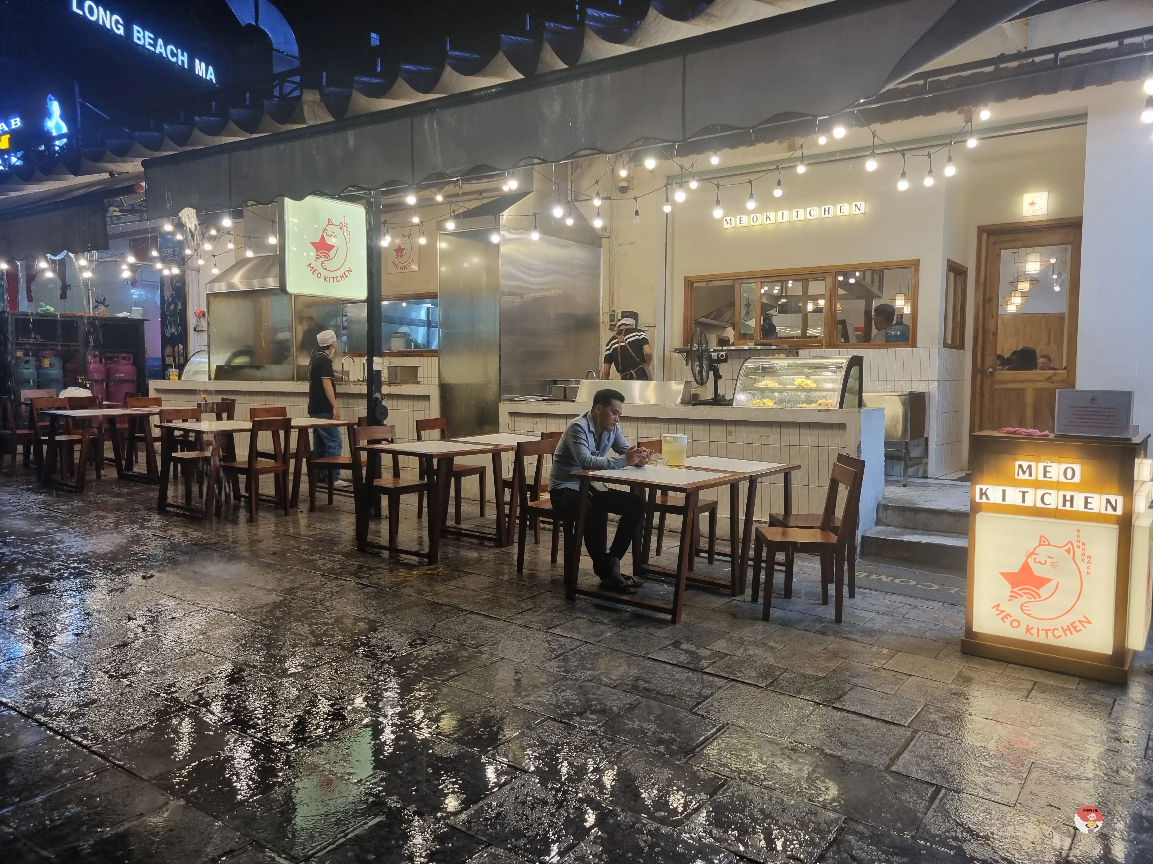 오는 거리의 야외 식당 ‘MEO KITCHEN’. 따뜻한 조명 아래 테이블과 의자가 놓여 있고, 한 남성이 앉아 있습니다.
