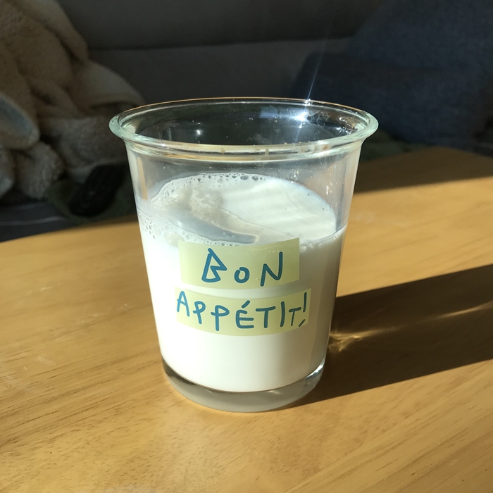 투명한 유리컵에 담긴 믈레코비타 갓밀크. &amp;#39;BON APP&Eacute;TIT!&amp;#39; 문구가 적힌 컵에 우유가 담겨 있으며 햇살이 비치는 테이블 위에 놓여 있다.