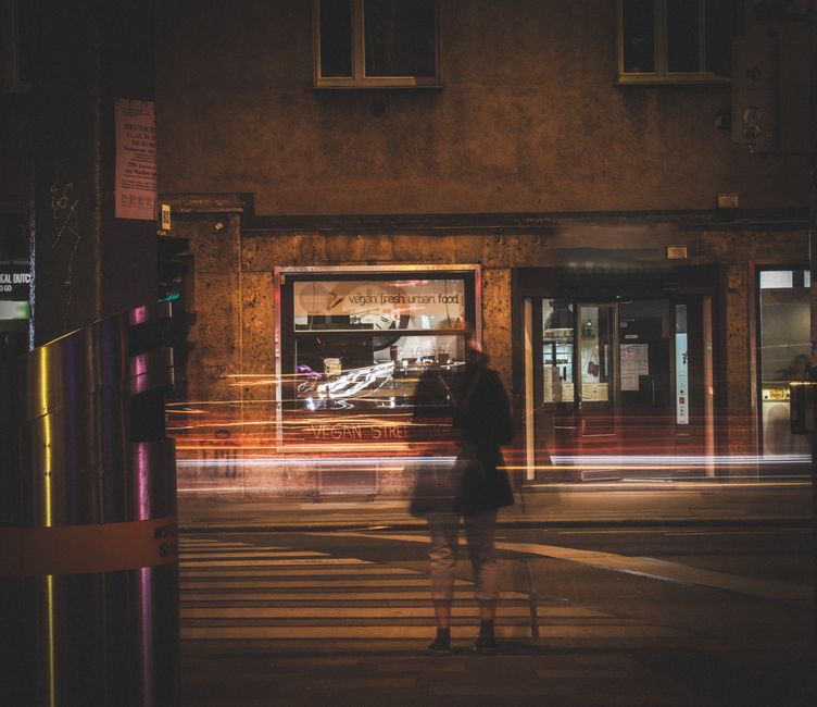 A ghostly figure at a pedestrian crossing in a dimly lit city with blurred motion lights.