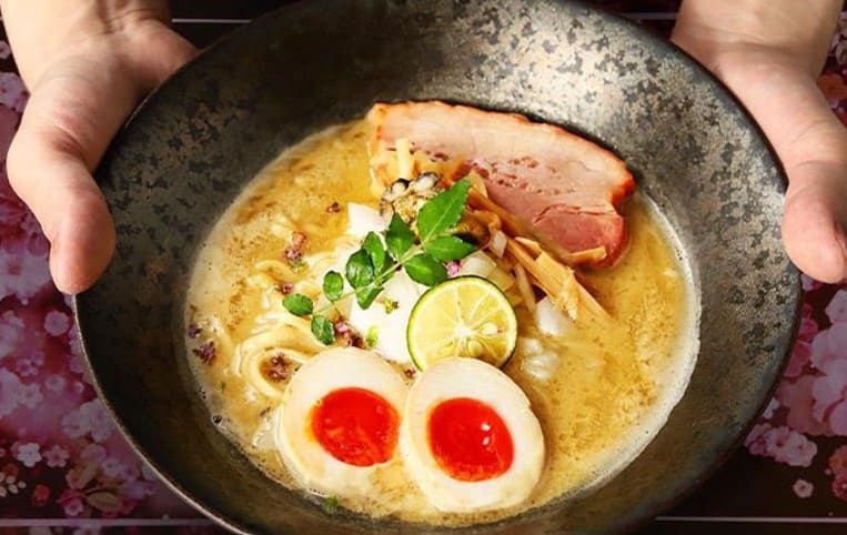 라면 그릇을 손으로 감싸고 있는 사진 라면에는 계란 반숙이 반으로 갈라져 있고 갈색 국물과 면 그리고 차슈가 올라가 있다