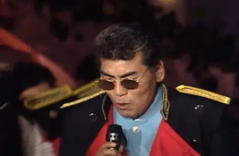 나훈아- 검은 옷 양쪽 어깨에 황금색 완장이 달린 옷을 입고 선그라스를 쓴채 노래하는 나훈아