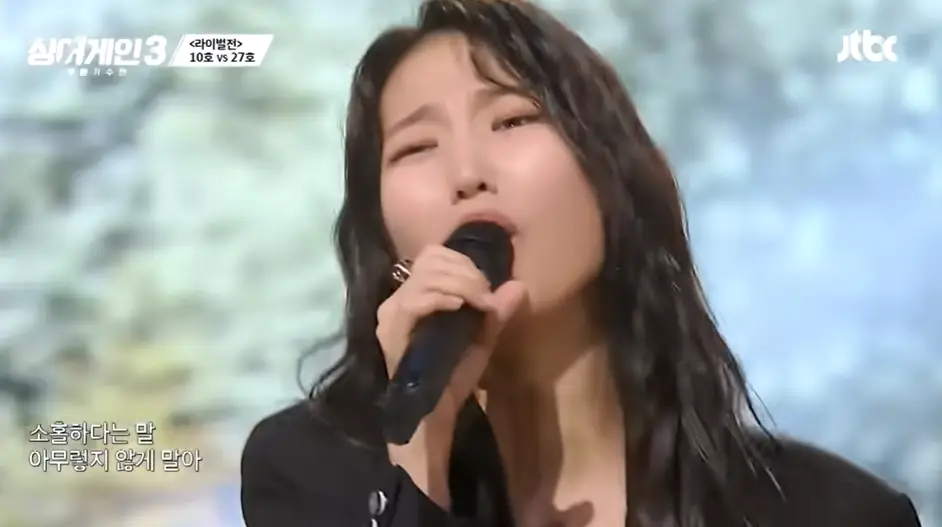 싱어게인3- 웨이브 진 검정색 긴머리로 오른손으로 마이크를 잡고 검정색 쟈켓을 정면을 바라보고 슬프게 노래하는 27호