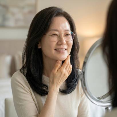 화장대 거울을 보며 손가락으로 탄력 있는 턱선을 살짝 만진 채 부드럽게 미소 짓는 50대 중년 여성