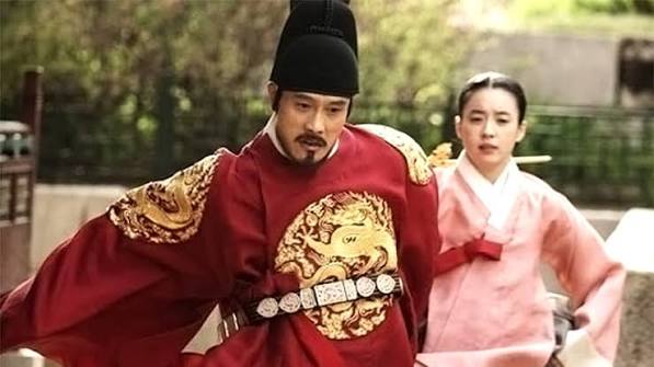 "영화 광해, 왕이 된 남자에서 붉은 용포를 입은 하선이 엉뚱하고 코믹한 표정으로 다급하게 뛰고 있고, 그 뒤에서 중전이 당황스러운 표정으로 왕의 손에 붙들려 함께 뛰고 있는 영화 속 한 장면"