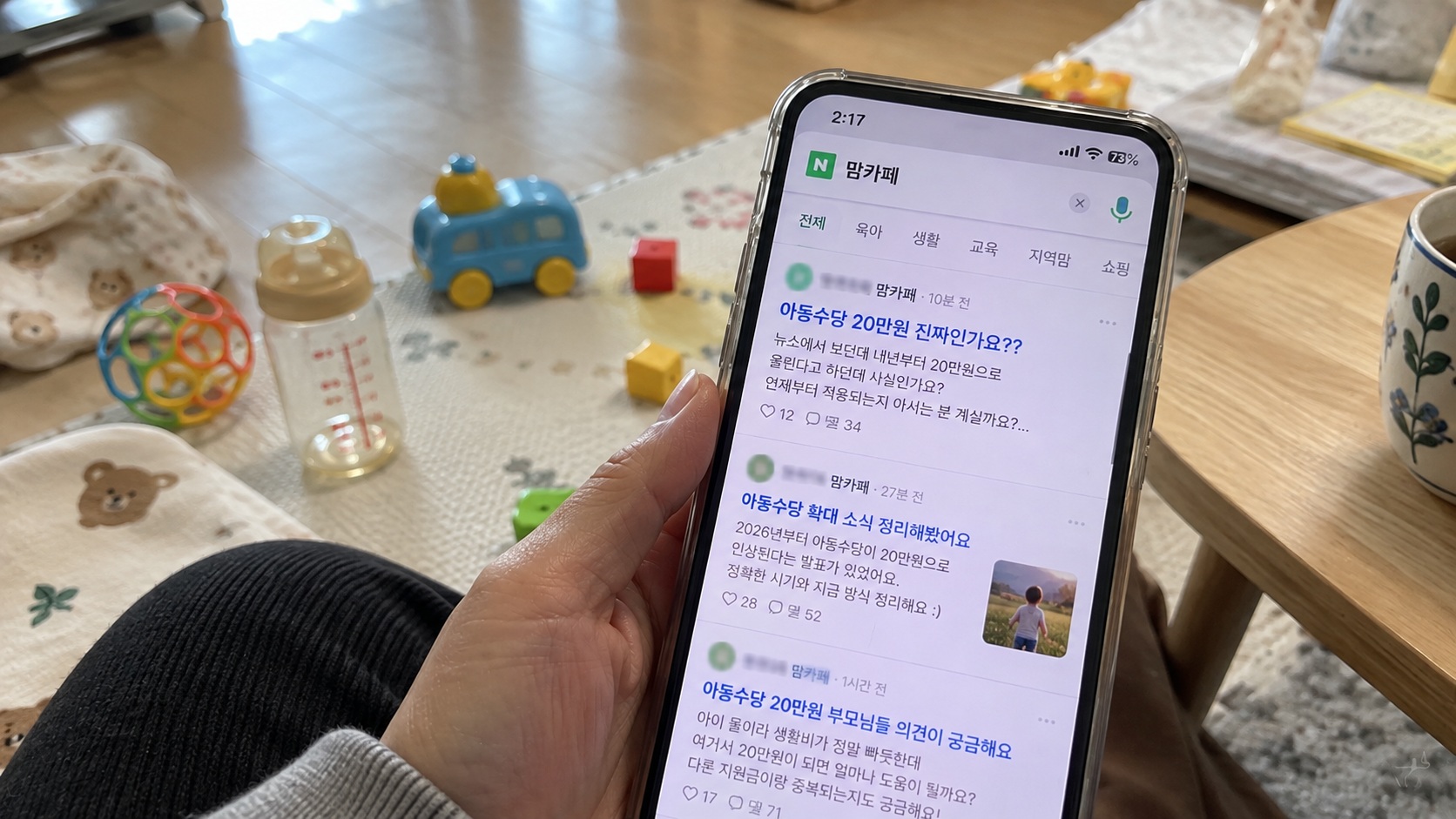 아기 장난감과 젖병이 놓인 거실에서 스마트폰으로 아동수당 20만원 확대 관련 맘카페 글을 확인하는 모습