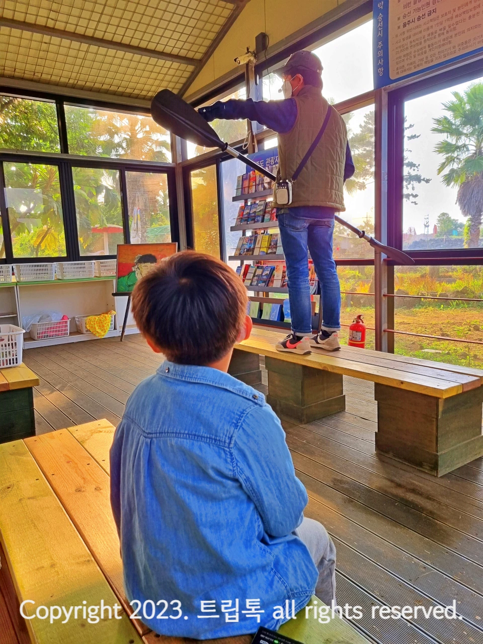 카양 노를 든 남자가 의자 위에서 설명을 하고 한 아이가 앉아서 듣고 있는 뒷모습이 보인다.