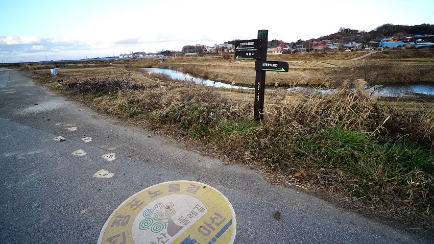 둑방길 바닥에 아산둘레길 길안내 스티커, 둑방길 우측에 둘레길 이정표, 우측에 실개천, 좌우에 잡초, 푸른 하늘, 들판 너머에 마을풍경,