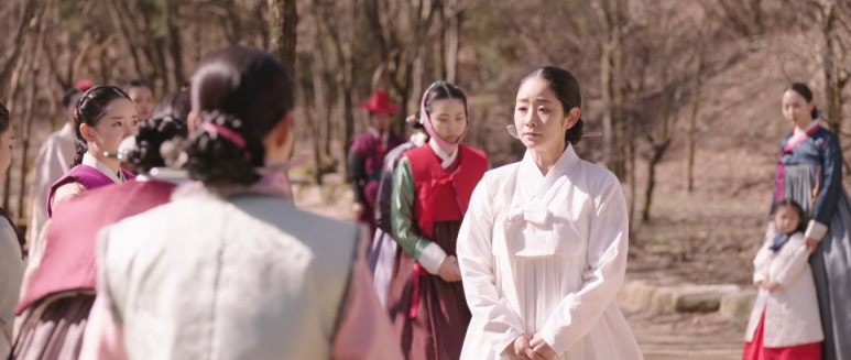 전통 의상을 입은 여성이 여러 사람 앞에서 담담한 표정으로 서 있는 모습