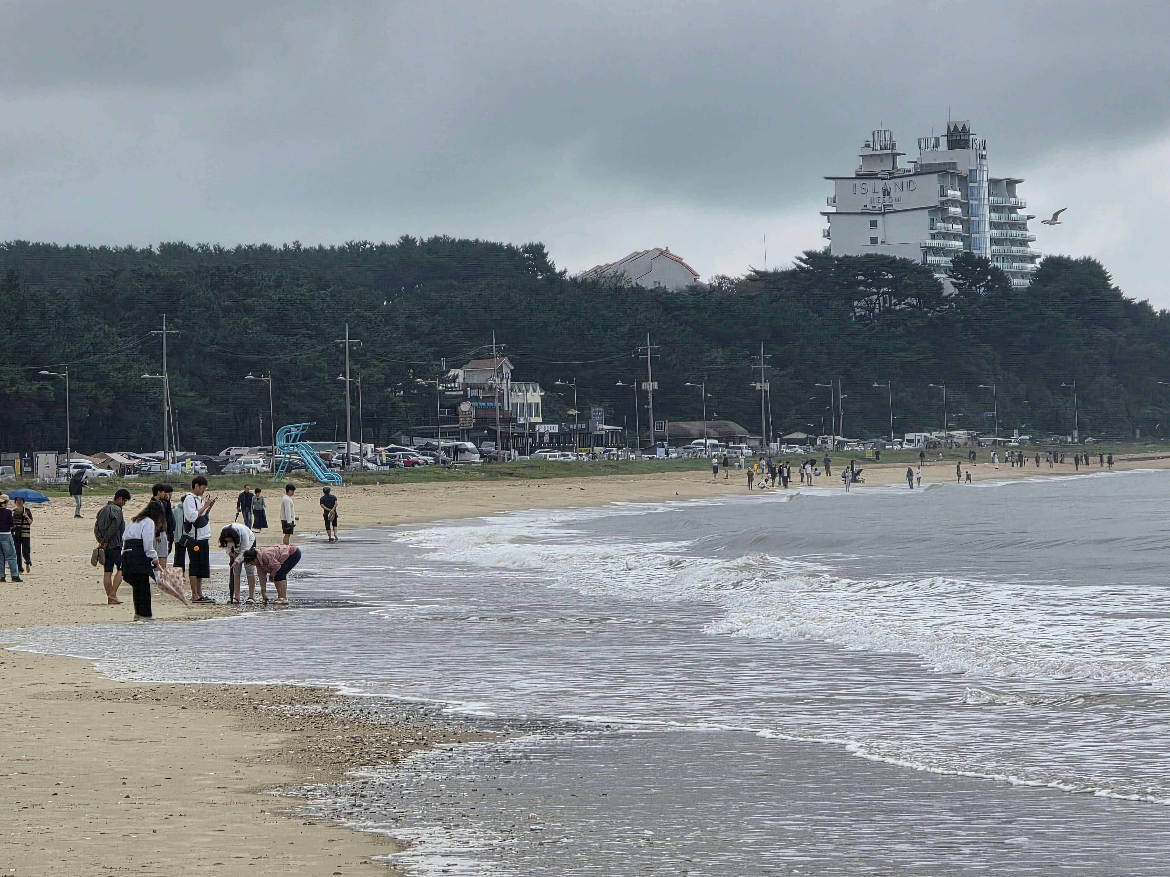 흐린 하늘 아래 안면도 해변 전경. 멀리 아일랜드 리조트 건물이 보이고 모래사장 위에 사람들이 여유롭게 걷고 있는 모습.