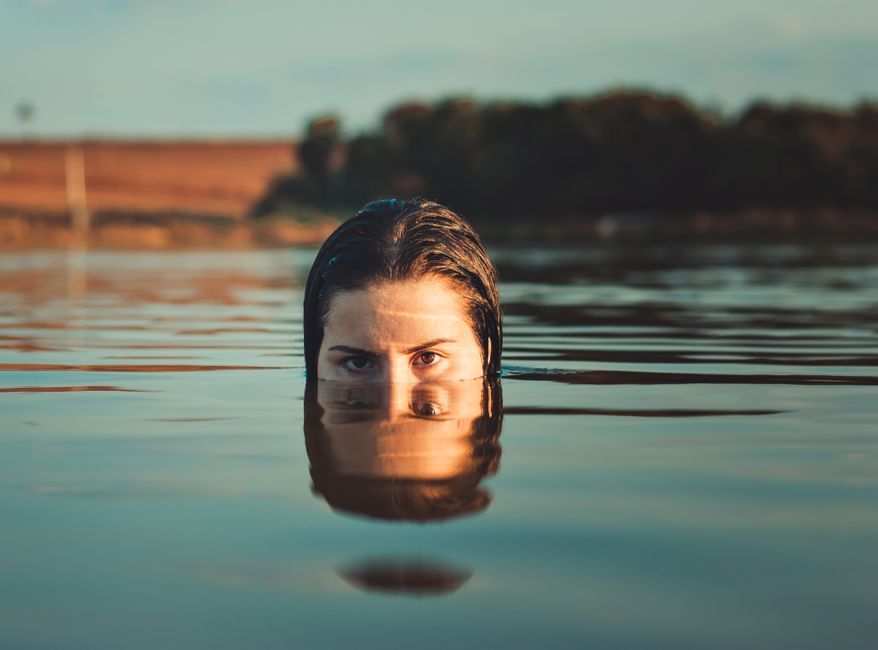 A woman gazes intensely while half-submerged in calm waters, creating a reflective scene.