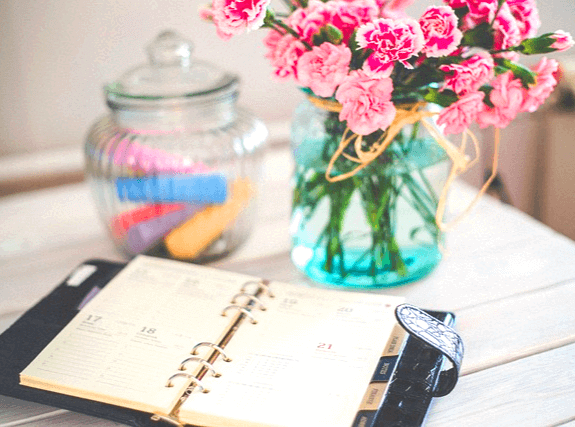 책상 위에 달력 수첩이 펼쳐져있고 분홍꽃이 가득한 파란 유리병 꽃병이 있다.