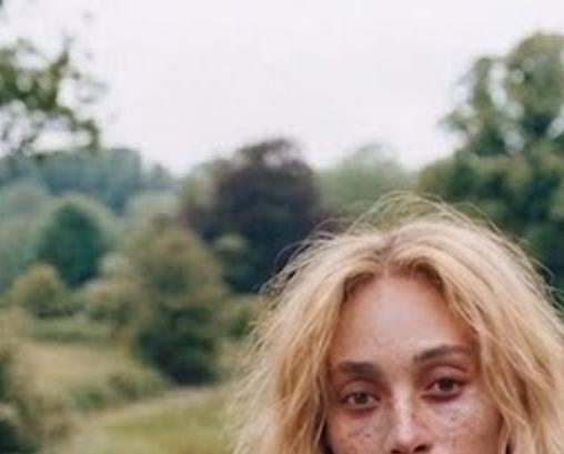 야외 초원 위에 앉아 있는 한 여성 모델이 베이지 톤 수트를 입고 카메라를 정면으로 바라보고 있으며, 자연 속 배경과 어우러진 차분하고 지속가능한 패션 화보 분위기의 이미지