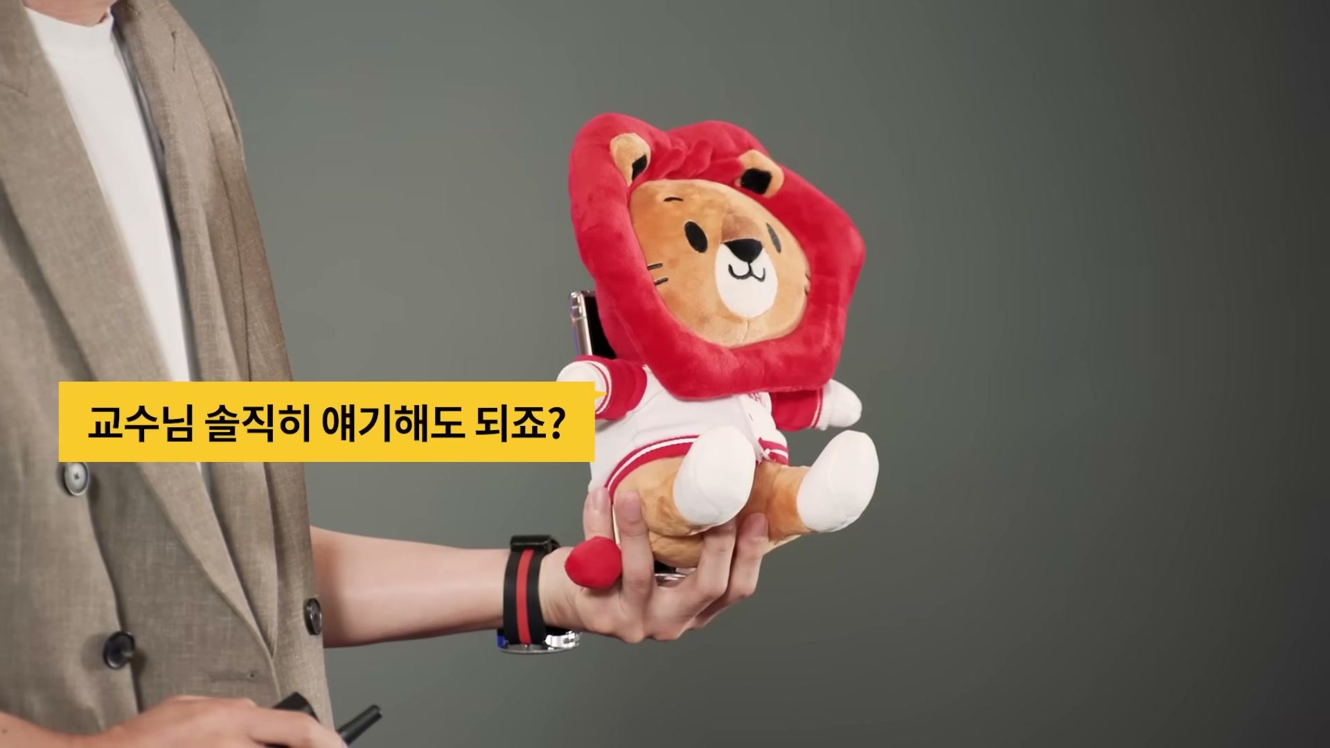빨간 갈기를 두른 사자 마스코트 쿠옹 인형 클로즈업과 함께 자막에 교수님 솔직히 얘기해도 되죠가 떠 있다.