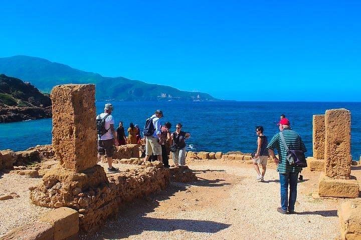 알제리 티파사의 고대 로마 유적지에서 관광객들이 유적 사이를 걷고 있다. 두 개의 거친 석주 사이로 보이는 배경에는 푸른 지중해와 멀리 펼쳐진 산맥이 보인다. 선명한 하늘과 햇살 아래 유적의 황토빛 질감이 뚜렷하게 드러난다.