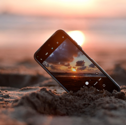 석양이 지는 바닷가 모래사장에 핸드폰이 45도 각도로 세워지고 화면은 카메라로 석양을 촬영 하는 이미지