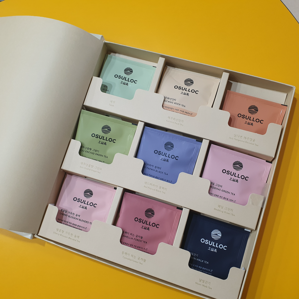 노란색 테이블 위에 오설록 시크릿 티 스토리 9종 세트가 놓여있다. 상자안에는 색이 다른 포장의 차가 보인다.