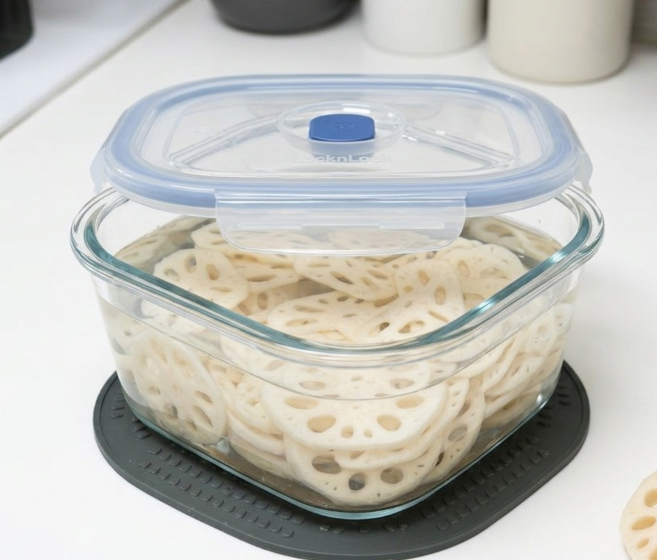 방 싱크대 위 그레이 실리콘 매트 위에 놓인, 물에 잠긴 슬라이스 연근이 가득 찬 투명 유리 밀폐 용기.