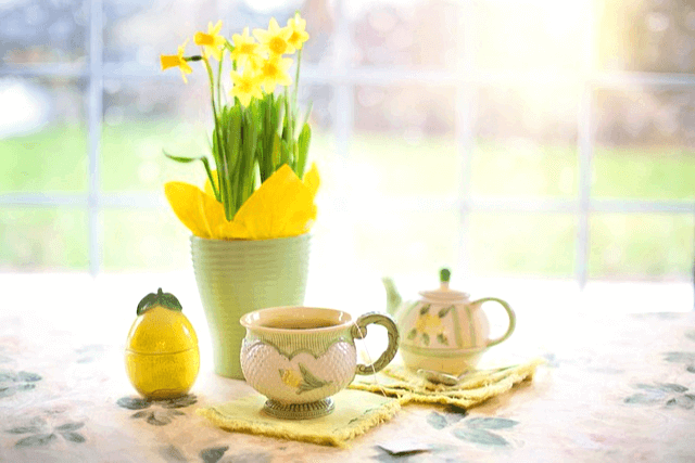 햇살이 비추는 거실에 하얀 식탁이 있고, 식탁 위에는 노란 수선화가 가득한 꽃병과 도자기 찻잔이 있다.