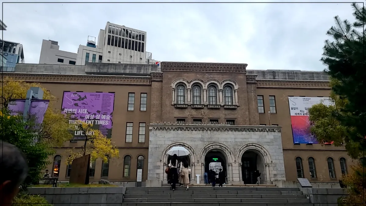 서울 시립 미술관 정면 모습, 벽에는 여성삶예술 간판, 미술관을 걸어들어가는 6명의 뒷모습, 하늘 양쪽에 푸른나무들