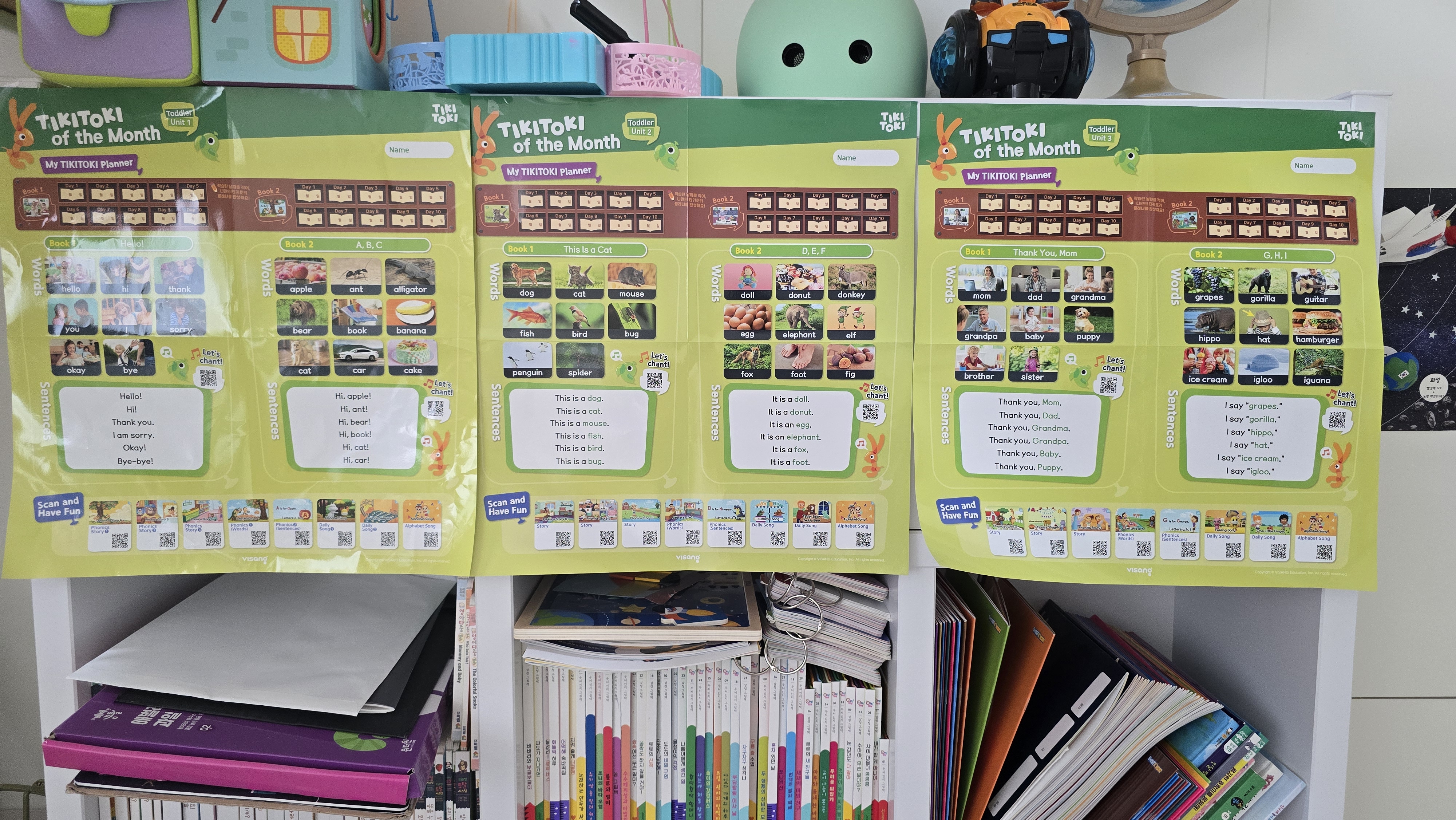 영유아 영어 단어와 문장이 정리된 티키토키 프로그램 포스터 3장이 벽에 나란히 붙어 있고, 아래 책장에는 다양한 영어 학습책과 어린이 책들이 정돈되어 있음.