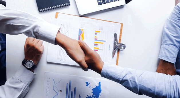 와이셔츠를 입은 남자 둘이 악수하는 장면이다. 책상에는 서류들이 있다.