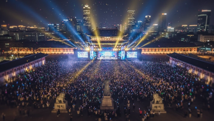 2026년 광화문광장에 모인 수많은 BTS 팬들이 콘서트 무대를 향해 환호하는 모습, 화려한 조명과 서울의 건축물들이 어우러져 활기찬 분위기를 연출함.