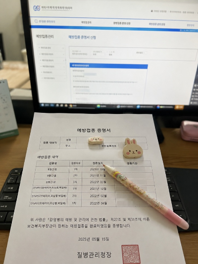 질병관리청 예방접종도우미 웹사이트 화면이 켜진 모니터 앞 키보드 위에 놓인 인쇄된 예방접종 증명서 종이 문서